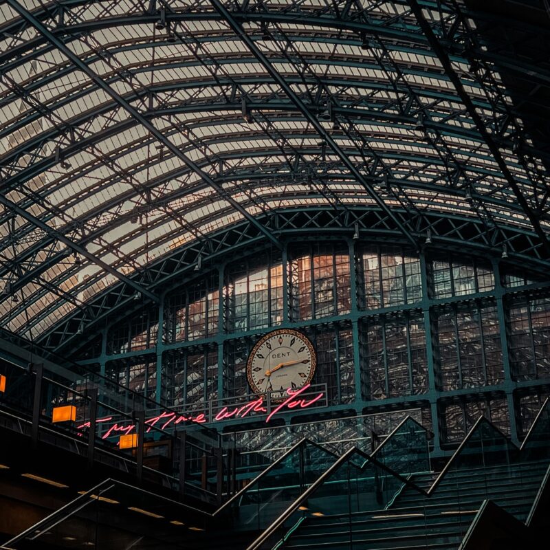 A large clock and neon sign, installed by a specialty contractor, are mounted on the glass wall of a train station under an arched, metal-framed roof.