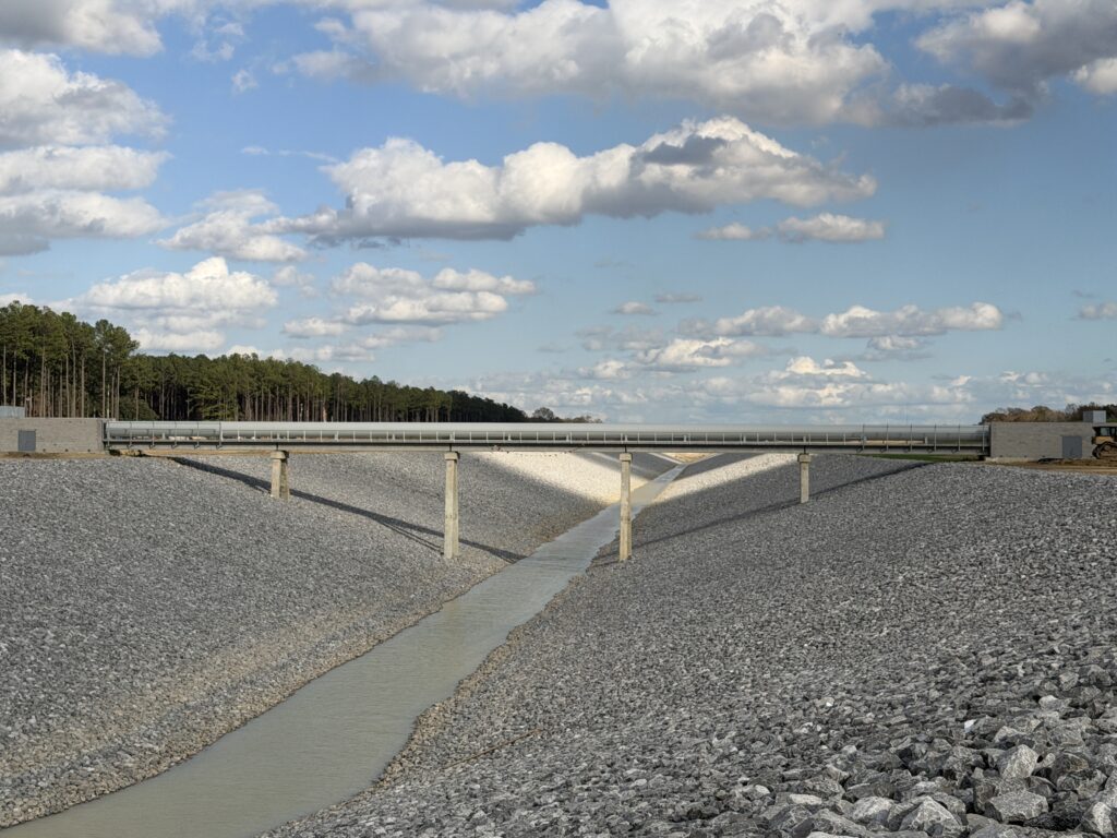 A concrete bridge by a specialty contractor spans a narrow, rocky drainage canal under a cloudy sky with trees in the background.